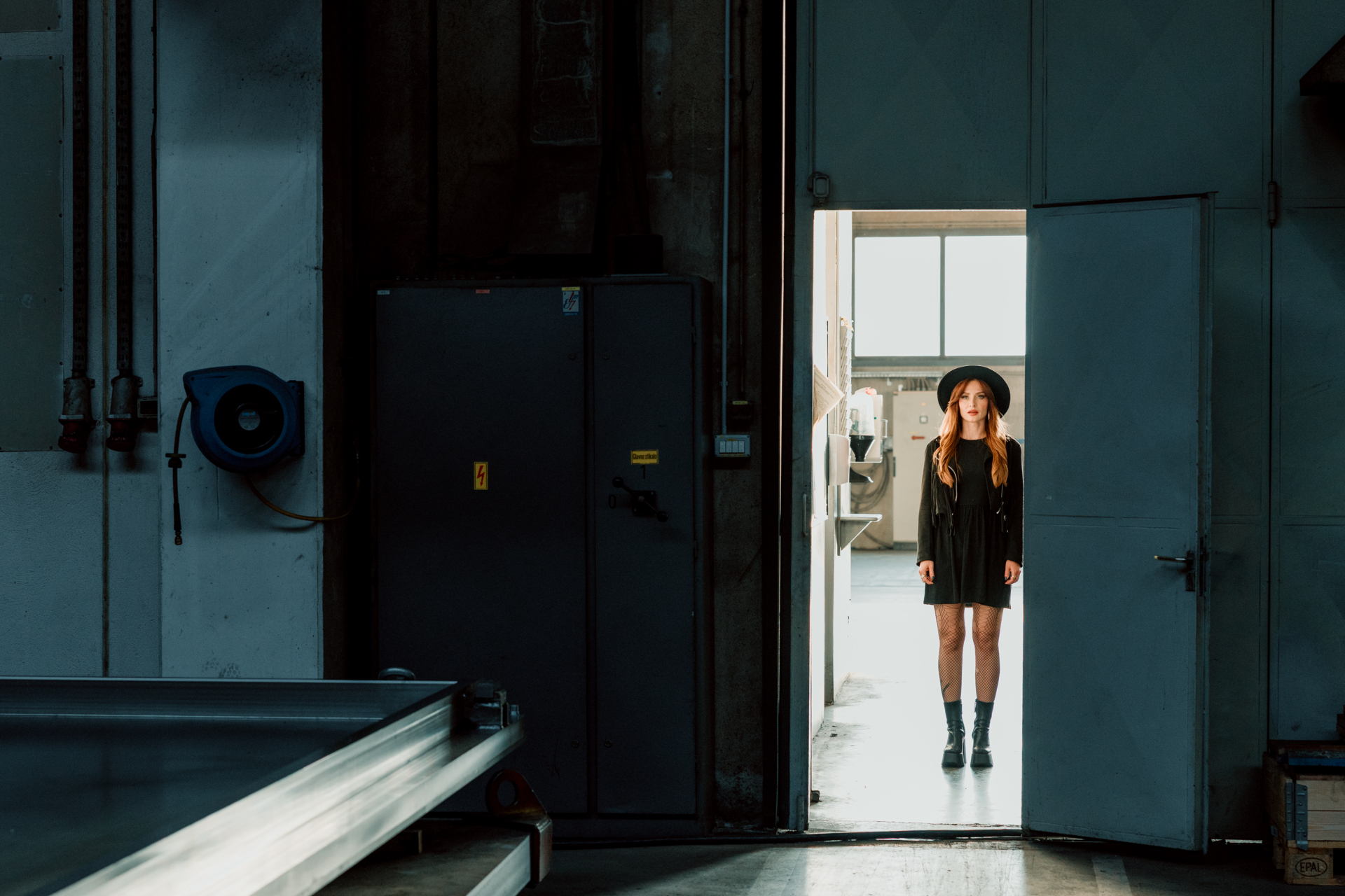 Kati standing in a backlit industrial doorway wearing a black dress and wide-brim hat.
